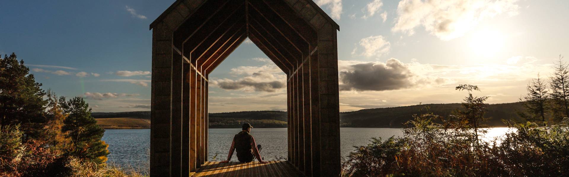 Robin’s Hut at Kielder Water, a wooden shelter framing a seated person looking out across the lake in warm evening light.