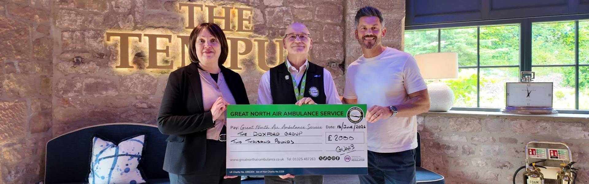 Three people stand inside The Tempus holding a presentation cheque for £2,000 from The Doxford Group to the Great North Air Ambulance Service. The group is photographed in front of a stone wall featuring an illuminated “The Tempus” sign.
